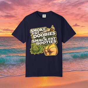 May include: A dark navy blue t-shirt featuring a graphic print with the text SMOKE FAT DOOBIES & SMACK FAT BOOTIES. The design includes a lit joint, a green cannabis bud, and a stylized illustration of buttocks wearing yellow underwear.