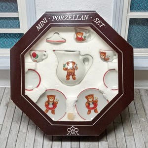 Op de afbeelding: Miniatuur porseleinen theeservies in een bruine achthoekige doos. De set bevat een theepot, kopjes, schotels en een melkkan, allemaal versierd met een teddybeerontwerp. De doos is gemarkeerd met "MINI - PORZELLAN - SET".
