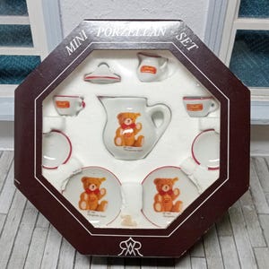 Op de afbeelding: Miniatuur porseleinen theeservies in een bruine achthoekige doos. De set bevat een theepot, melkkan, suikerpot, twee kopjes en twee schoteltjes. Elk stuk is wit met een rode rand en heeft een teddybeer illustratie. De doos leest "Mini Porzellan Set".