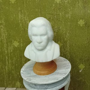 Könnte beinhalten: Eine kleine, weiße Marmorbüste eines Mannes mit langem Haar und Hemdkragen. Die Büste ist auf einem Holzsockel montiert, der auf einem kleinen, runden, dekorativen Tisch mit einem verwitterten, grau-weißen Anstrich steht.