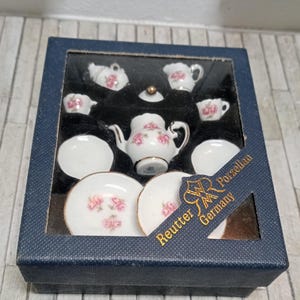 Może przedstawiać: Miniaturowy porcelanowy zestaw do herbaty w granatowym pudełku. Zestaw zawiera czajniczek, filiżanki, spodki i śmietankę, wszystkie zdobione różowymi różami i złotymi obwódkami. Pudełko ma przezroczyste okienko i etykietę z napisem "Reutter Porzellan Germany."