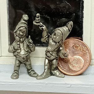 Op de afbeelding: Twee kleine, zilverkleurige kabouterbeeldjes. Eén kabouter wijst met een vinger, de andere houdt een klein voorwerp omhoog. Een euromunt ligt naast de kabouters. De kabouters staan op een wit oppervlak.