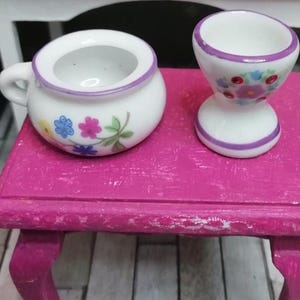 Op de afbeelding: Twee miniatuur porseleinen potten met bloemenmotieven. De grotere pot heeft een handvat en een paarse rand. De kleinere pot is een pot met een paarse rand en een bloemenmotief.