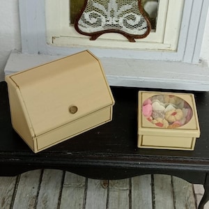 May include: Two miniature beige boxes with gold trim are displayed on a dark surface. One box is closed with a small gold button, and the other has a clear lid showing colourful miniature treats. The boxes are part of a dollhouse collection.