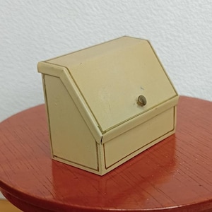 May include: A small, cream-coloured box with a hinged lid and a brass knob. The box has gold trim and is set on a red table. The box is likely a miniature storage container or decorative item.