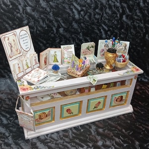 Op de afbeelding: Een miniatuur naaiwinkel display met een wit en goud geschilderde toonbank. De toonbank heeft lades met engelen- en bloemmotieven. De display bevat miniatuur naaipatronen, scharen en een mandje met garen. Het bordje luidt "McCalls Patterns".