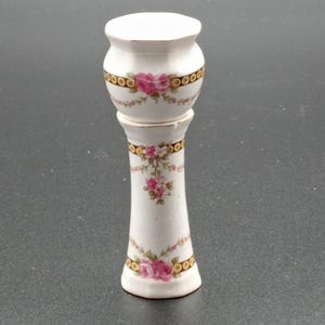 Könnte beinhalten: Ein weißer Keramiksockel mit floralem Design. Der Sockel hat eine runde Oberseite und eine hohe, schlanke Basis. Das Blumenmuster zeigt rosa Rosen und grüne Blätter.