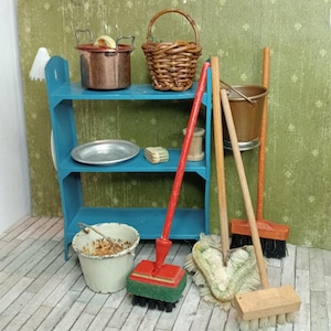 Op de afbeelding: Miniatuur schoonmaakartikelen en keukengerei voor een poppenhuis. Inclusief een blauw rek, koperen pot, gevlochten mand, metalen emmer, bezems en een dweil. De items staan op een vloer met houtlook tegen een groene behangachtergrond.