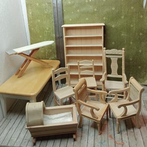Op de afbeelding: Een verzameling onafgewerkte houten poppenhuismeubels, waaronder een boekenplank, stoelen, een tafel, een strijkplank en een wieg. De stukken zijn licht van kleur en lijken van natuurlijk hout te zijn, met zichtbare nerfdetails. De achtergrond is groen.