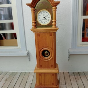 Könnte beinhalten: Eine Miniatur-Standuhr aus hellem Holz. Das Zifferblatt der Uhr zeigt römische Ziffern und schwarze Zeiger. Die Uhr hat eine dekorative Bogenoberseite und eine runde Öffnung mit einem goldenen Pendel. Etwa 25 cm hoch.