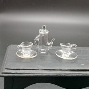 Op de afbeelding: Een miniatuur glazen theeset met een heldere theepot en twee kopjes en schoteltjes. De set staat op een zwart houten oppervlak.