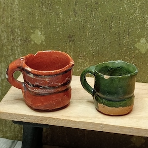 Op de afbeelding: Twee kleine keramische bekers, een is oranje met zwarte strepen en de andere is groen met een bruine rand. De bekers staan op een houten plank.