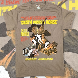 May include: A brown t-shirt with a vintage movie poster design. The poster features a scene from the film "Death Rides a Horse" with the tagline "This is Revenge... and there's nothing sweet about it." The poster also includes the names of the actors, Lee Van Cleef and John Phillip Law.