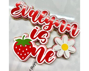 May include: A red and white cake topper with the words "Evelyn is one" and a strawberry and daisy design.