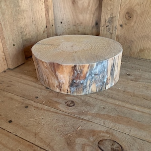 May include: A round, natural wood slice with a light brown top and a rough, textured edge. The side of the wood slice has a mix of brown, white, and gray tones. The wood slice is a rustic decorative item.
