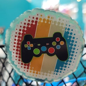 May include: A blue and white circular badge with a retro-style rainbow design and a black video game controller graphic. The badge has a silver chain attached.