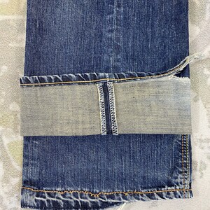 May include: A pair of dark blue denim jeans with a folded cuff revealing a light grey interior. The cuff features a visible vertical white selvedge seam with white stitching. The hem is finished with thick orange thread stitching along the edge.