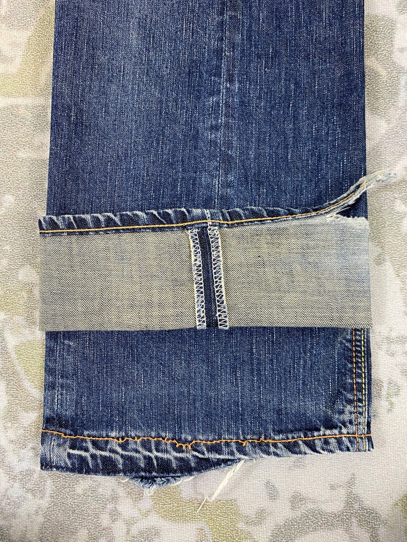 May include: A pair of dark blue denim jeans with a folded cuff revealing a light grey interior. The cuff features a visible vertical white selvedge seam with white stitching. The hem is finished with thick orange thread stitching along the edge.