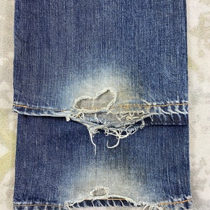 May include: A pair of blue denim jeans featuring significant wear and fraying at the bottom hems. The fabric shows a faded wash with visible distressed areas, loose white threads, and worn edges along the cuffs of the pant legs.
