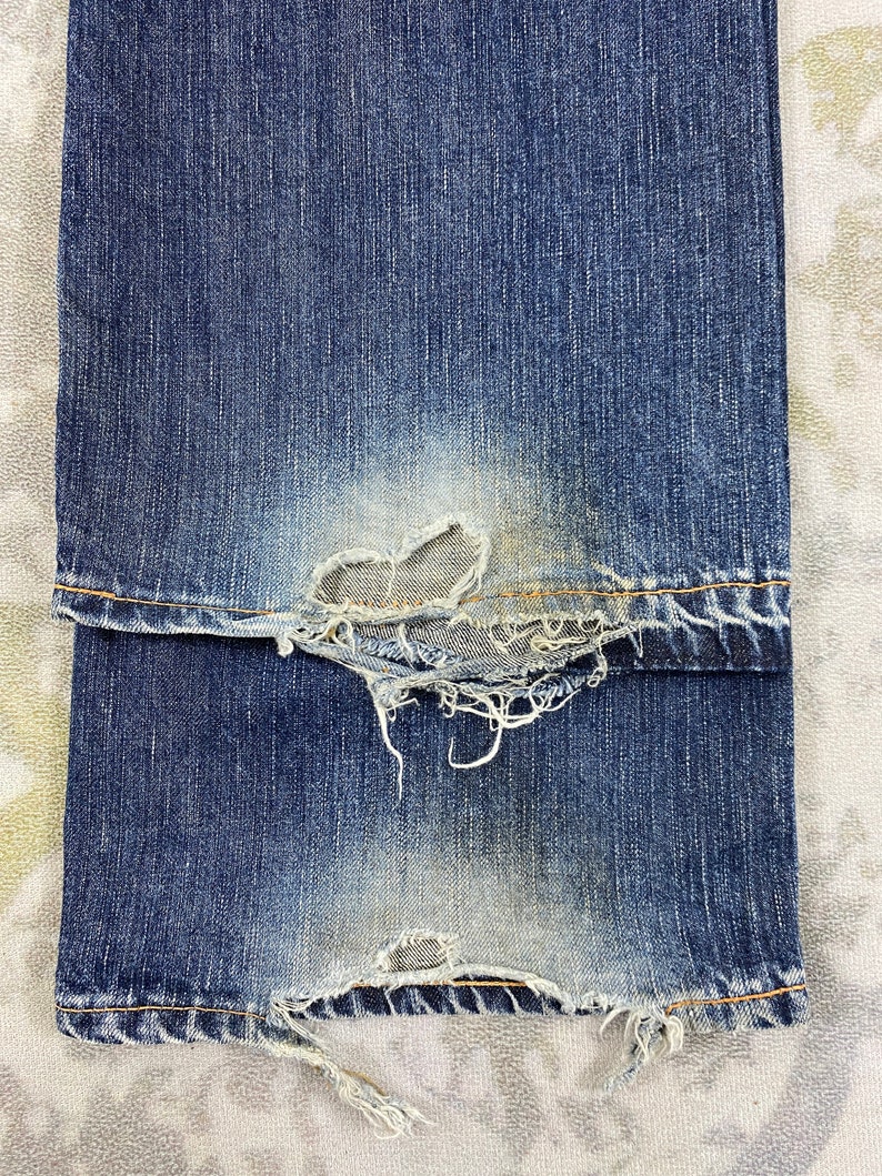 May include: A pair of blue denim jeans featuring significant wear and fraying at the bottom hems. The fabric shows a faded wash with visible distressed areas, loose white threads, and worn edges along the cuffs of the pant legs.