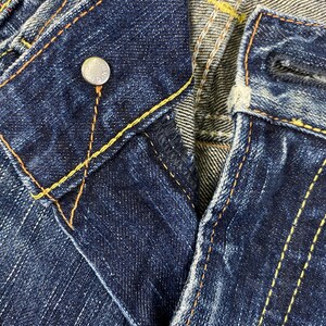 May include: Dark blue denim jeans featuring a metal button closure with embossed text, a button fly, and prominent orange and yellow contrast stitching along the seams and waistband. The fabric displays a textured, worn-in wash finish.