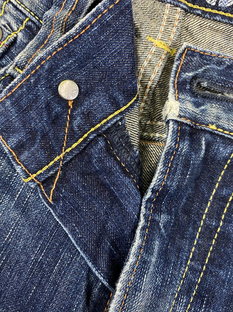 May include: Dark blue denim jeans featuring a metal button closure with embossed text, a button fly, and prominent orange and yellow contrast stitching along the seams and waistband. The fabric displays a textured, worn-in wash finish.