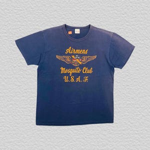 May include: Navy blue t-shirt with a crew neck. The shirt features a graphic with the text "Airmens Mosquito Club U.S.A.F." in yellow and orange. The graphic includes a stylized mosquito with wings. The shirt has short sleeves.