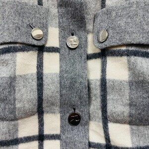 K&ouml;nnte beinhalten: Nahaufnahme einer grau-wei&szlig; karierten Jacke mit silbernen Kn&ouml;pfen. Die Jacke hat zwei Taschen und einen Knopfverschluss vorne. Die Kn&ouml;pfe haben ein kreisf&ouml;rmiges Design.