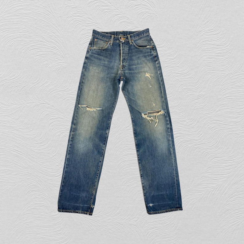 May include: Faded blue denim jeans with distressed details, including ripped knees and a button fly. The jeans have a classic five-pocket design and a straight leg fit. The denim has a worn appearance.