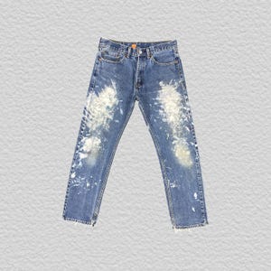 Puede incluir: Pantalones vaqueros de mezclilla azul medio con un efecto de salpicaduras de pintura blanca en la parte delantera de ambas piernas. Estos vaqueros incluyen un cierre de botones estándar, cinco bolsillos y un corte recto con costuras visibles.