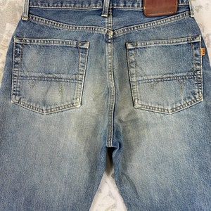 May include: Back view of blue denim jeans with two back pockets and a brown leather patch. The jeans have a faded wash and a classic five-pocket design. The stitching is visible, and the jeans appear to be well-worn.