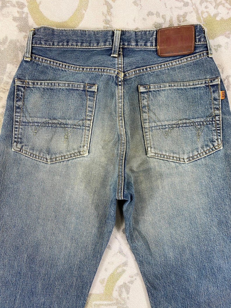 May include: Back view of blue denim jeans with two back pockets and a brown leather patch. The jeans have a faded wash and a classic five-pocket design. The stitching is visible, and the jeans appear to be well-worn.