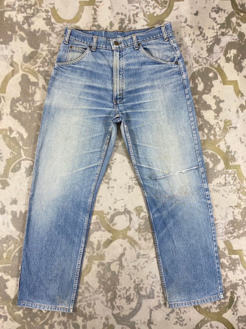 K&ouml;nnte beinhalten: Hellblaue Jeans aus Denim mit verwaschenem Look und abgenutztem Aussehen. Die Jeans haben ein klassisches F&uuml;nf-Taschen-Design, einen Knopf- und Rei&szlig;verschluss und ein gerades Bein. Auf dem rechten Knie befindet sich ein Flicken. Die Jeans liegen flach auf einer gemusterten Oberfl&auml;che.