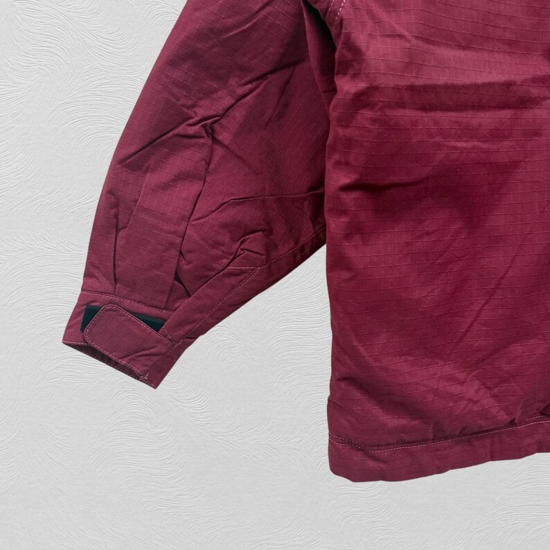 May include: A close-up of a maroon jacket sleeve. The sleeve has a textured, ribbed pattern and a black velcro cuff closure. The jacket appears to be made of a lightweight material, suitable for layering. The background is a neutral, textured surface.