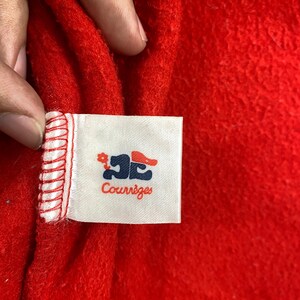 K&ouml;nnte beinhalten: Nahaufnahme eines roten Stoffes mit einem wei&szlig;en Etikett. Das Etikett zeigt ein Logo mit einer Blume, einem Schuh und dem Wort "Courr&egrave;ges" in Rot. Das Etikett ist mit roten N&auml;hten auf den Stoff gen&auml;ht.