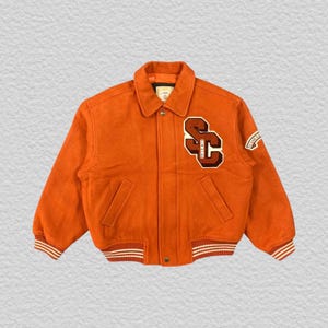 May include: An orange varsity jacket featuring a fold-down collar, long sleeves, and two front pockets. The left chest displays a large brown and cream patch with the letters SC and the text SIM CLUB. A small patch is on the right sleeve.