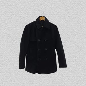 May include: A black double-breasted pea coat featuring a wide collar, long sleeves, and six dark buttons arranged in two vertical rows on the front. The coat is displayed on a wooden hanger, showcasing a structured design with a straight hemline.