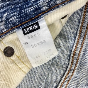 May include: Close-up of a pair of blue denim jeans with a white tag. The tag reads "EDWIN" and includes the size 30x33. The jeans have brown stitching and metal rivets.