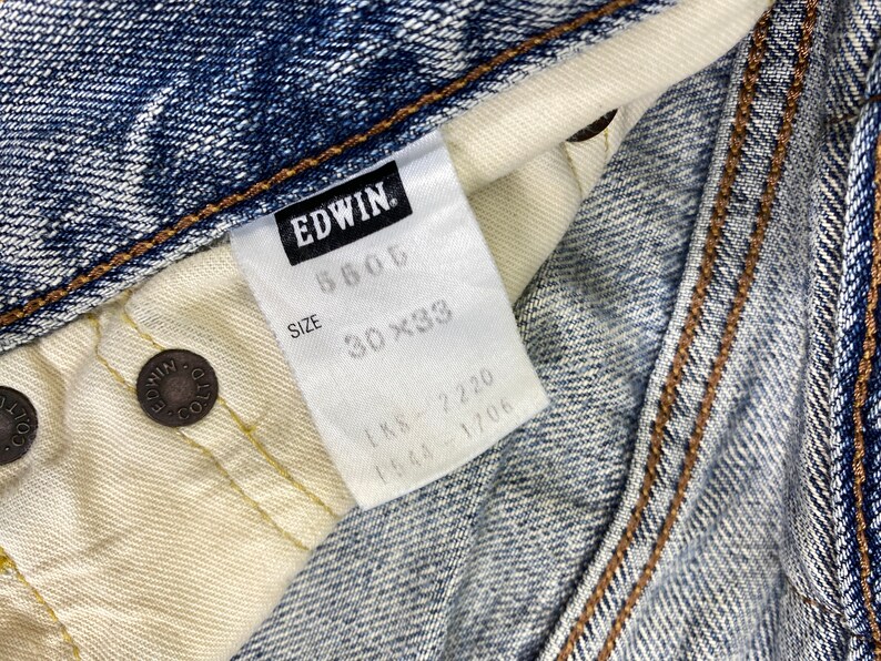 May include: Close-up of a pair of blue denim jeans with a white tag. The tag reads "EDWIN" and includes the size 30x33. The jeans have brown stitching and metal rivets.