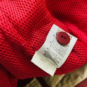 May include: Close-up of a red mesh fabric with a red button and a white tag. The tag reads "MADE IN VIETNAM" in black text. The fabric appears to be part of a garment.