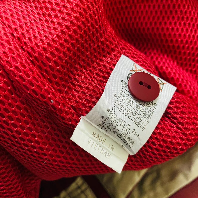 May include: Close-up of a red mesh fabric with a red button and a white tag. The tag reads "MADE IN VIETNAM" in black text. The fabric appears to be part of a garment.