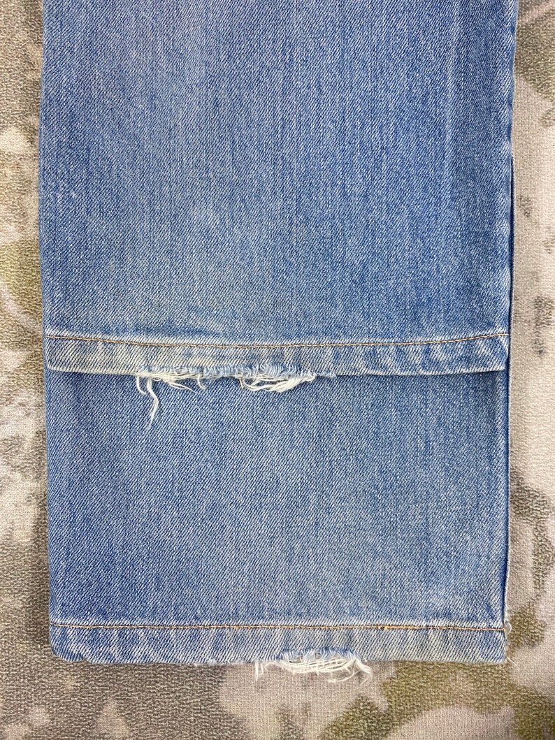 K&ouml;nnte beinhalten: Nahaufnahme einer hellblauen Jeans. Der Saum der Jeans ist ausgefranst und abgenutzt, mit sichtbaren F&auml;den. Der Stoff hat eine klassische Denim-Textur und -Farbe.