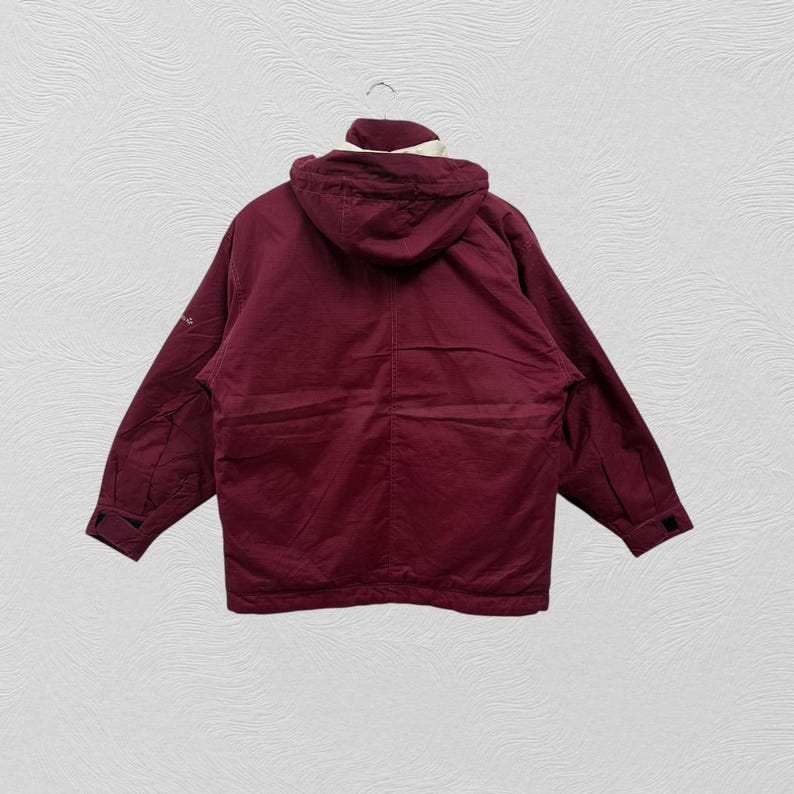 May include: A maroon hooded jacket with a white and cream-colored lining around the hood. The jacket has a full zipper, two pockets, and black cuffs. The jacket is hanging on a metal hanger against a white textured background.