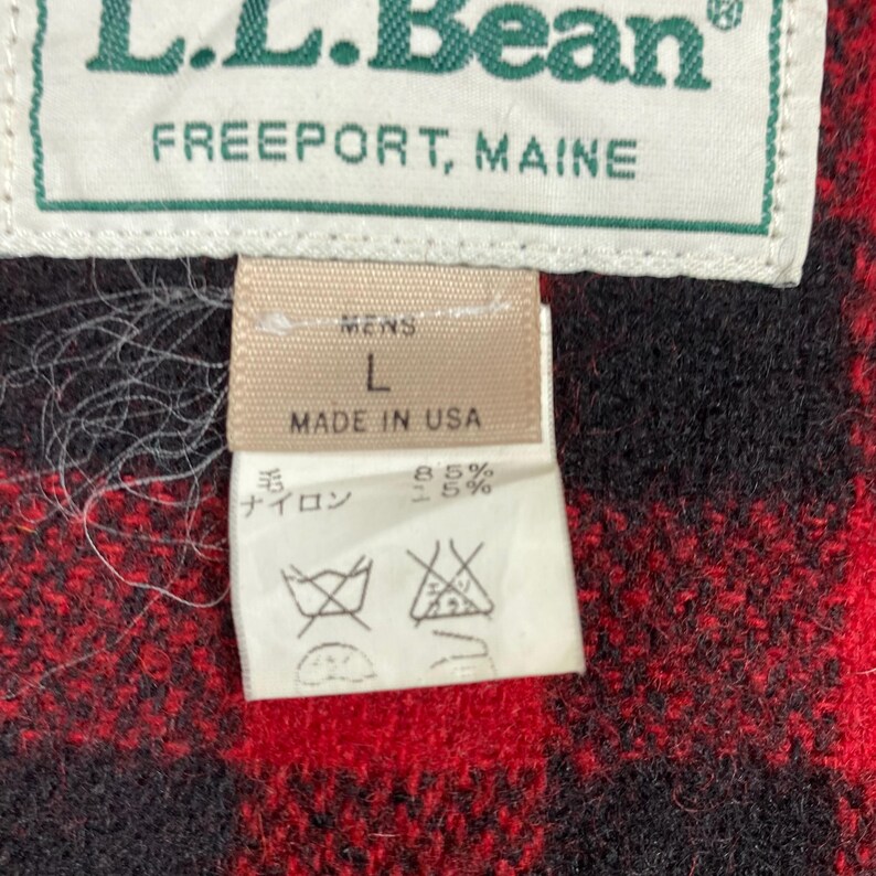 Op de afbeelding: Close-up van een label van een L.L.Bean kledingstuk. Het label luidt "L.L.Bean FREEPORT, MAINE" in groen en wit. Daaronder staat een tan label met "MENS L MADE IN USA". Een kleiner label toont wasvoorschriften en de samenstelling van de stof.