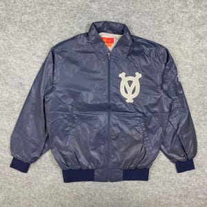 May include: A navy blue jacket featuring a cream-colored emblem. The jacket has a collared neckline, a zip closure, and dark blue ribbed cuffs and hem. The jacket has a slight sheen.