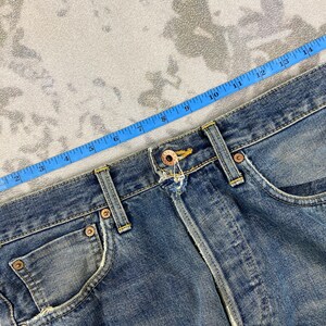May include: Close-up of blue denim jeans with a measuring tape laid across the waistband. The jeans have a classic five-pocket design, with visible stitching and metal rivets. The denim has a faded, worn appearance.