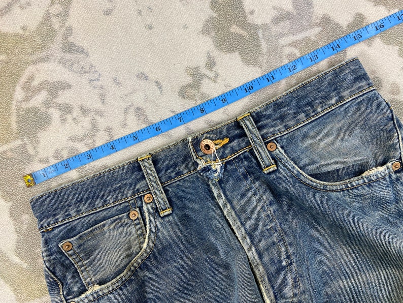 May include: Close-up of blue denim jeans with a measuring tape laid across the waistband. The jeans have a classic five-pocket design, with visible stitching and metal rivets. The denim has a faded, worn appearance.