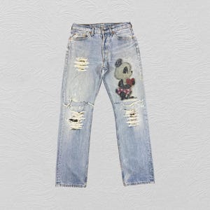 May include: Light blue denim jeans featuring a distressed, ripped finish on the legs. The front of the jeans displays a graphic patch of a cartoon panda holding a red heart. The pants include a standard button fly closure and belt loops.