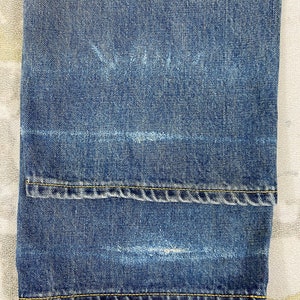 May include: Close-up of blue denim jeans with a folded hem. The jeans have a faded appearance with visible wear marks. Yellow stitching is present along the hem and seams, adding contrast to the blue fabric. The jeans are folded at the bottom.