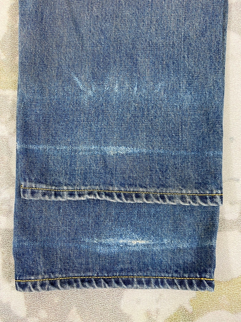 May include: Close-up of blue denim jeans with a folded hem. The jeans have a faded appearance with visible wear marks. Yellow stitching is present along the hem and seams, adding contrast to the blue fabric. The jeans are folded at the bottom.